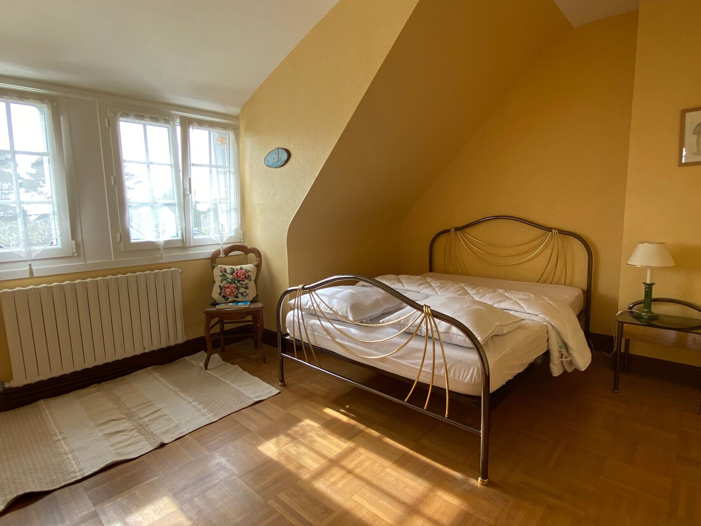 A bedroom with a sloped ceiling, a bed with metal headboard, a chair with a floral cushion, a bedside table with a lamp, and a window with curtains.