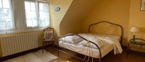 A bedroom with a sloped ceiling, a bed with metal headboard, a chair with a floral cushion, a bedside table with a lamp, and a window with curtains.