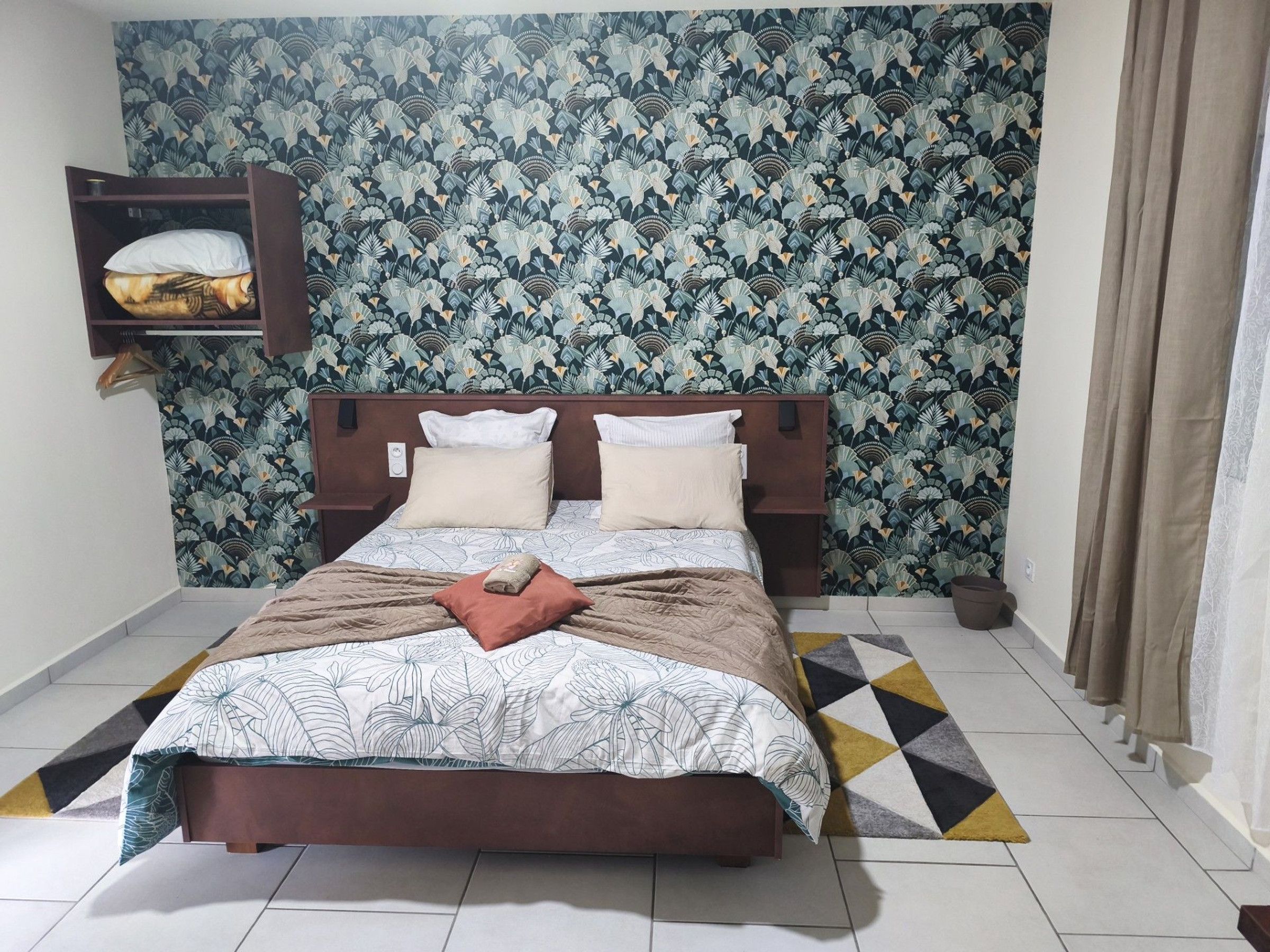 A neatly made bed with a patterned bedspread, a wooden headboard, and a small shelf with a folded towel.