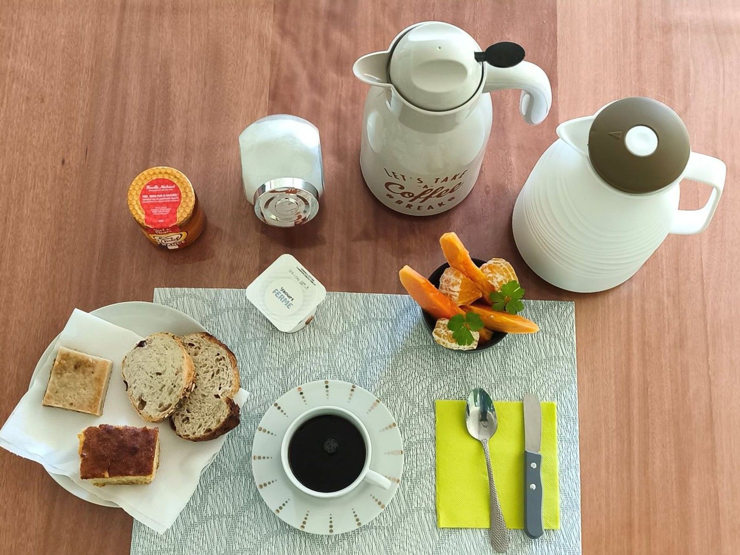 A breakfast setup with a coffee pot, teapot, and teacup, along with bread, butter, jam, and orange slices on a table.