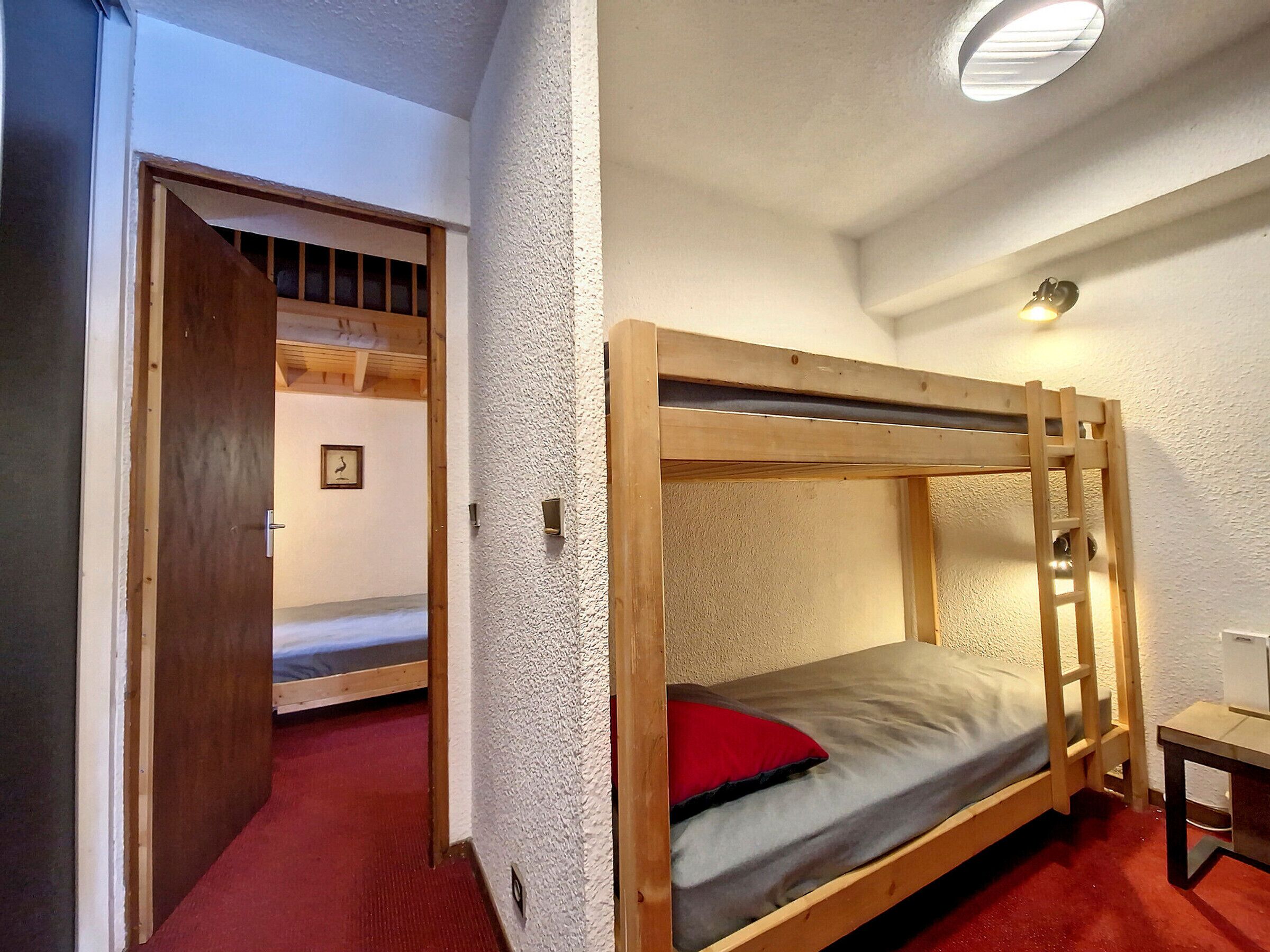 Une chambre avec un lit superposé, un tapis rouge, une porte en bois et une petite table.