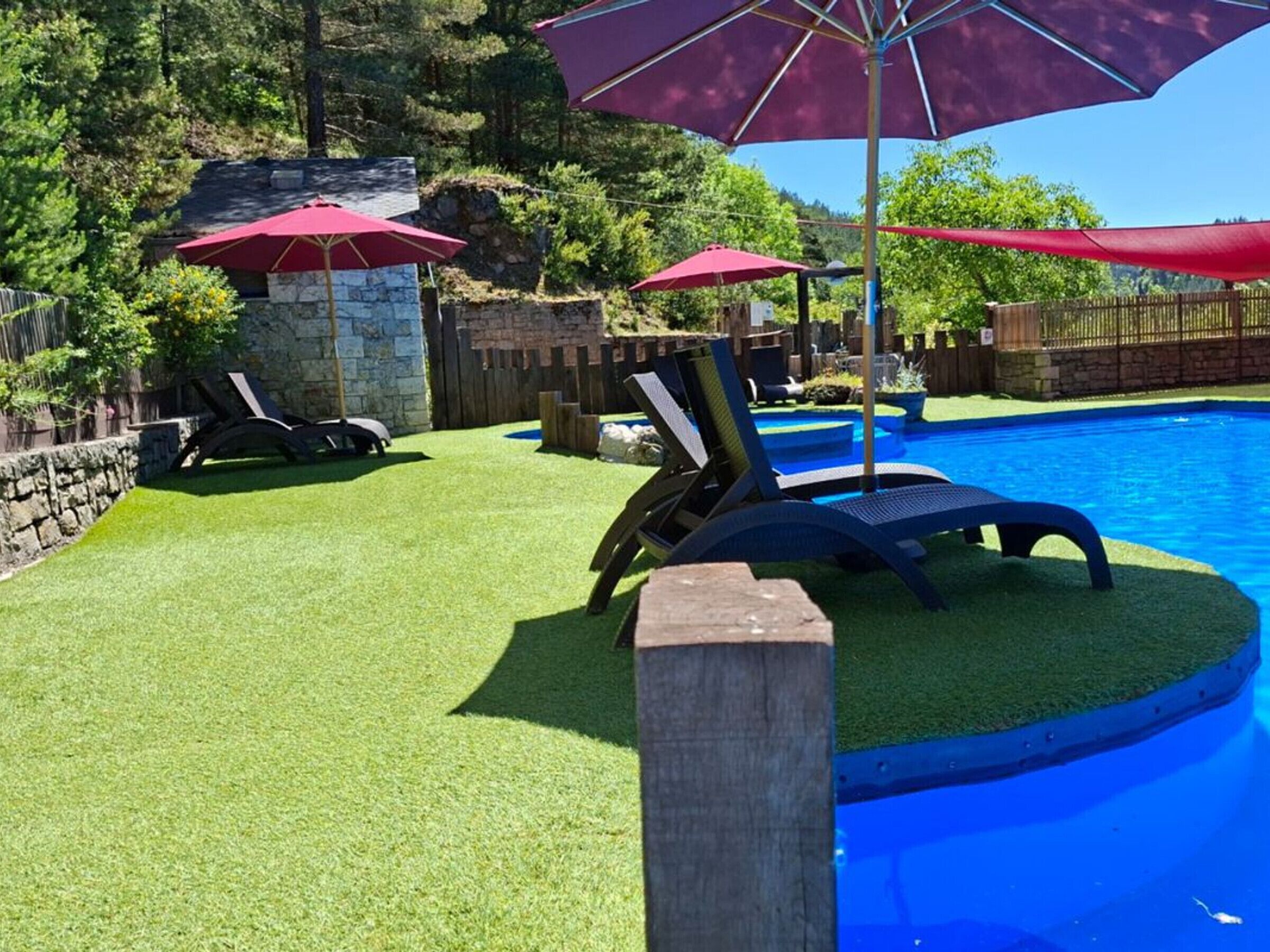 A backyard with a swimming pool, surrounded by a stone wall and wooden fence, with lounge chairs and umbrellas.