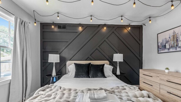 A modern bedroom with a large bed, two bedside tables with lamps, a wooden dresser, and a cityscape painting on the wall.
