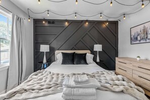 Une chambre moderne avec un grand lit, deux tables de chevet avec des lampes, une commode en bois et un tableau représentant un paysage urbain accroché au mur.
