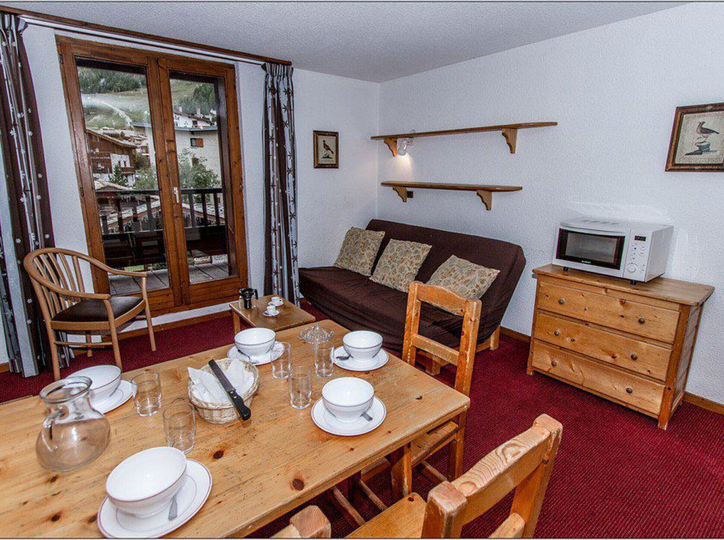 Una habitación acogedora con una mesa de comedor de madera para cuatro personas, un sofá marrón, un armario de madera y un televisor sobre un mueble de madera.