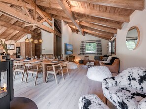 Un loft spacieux avec de hauts plafonds, des poutres en bois, un coin repas avec une table dressée pour six personnes, un coin salon confortable avec un canapé et un panier pour chien, ainsi qu’une grande fenêtre avec des rideaux.