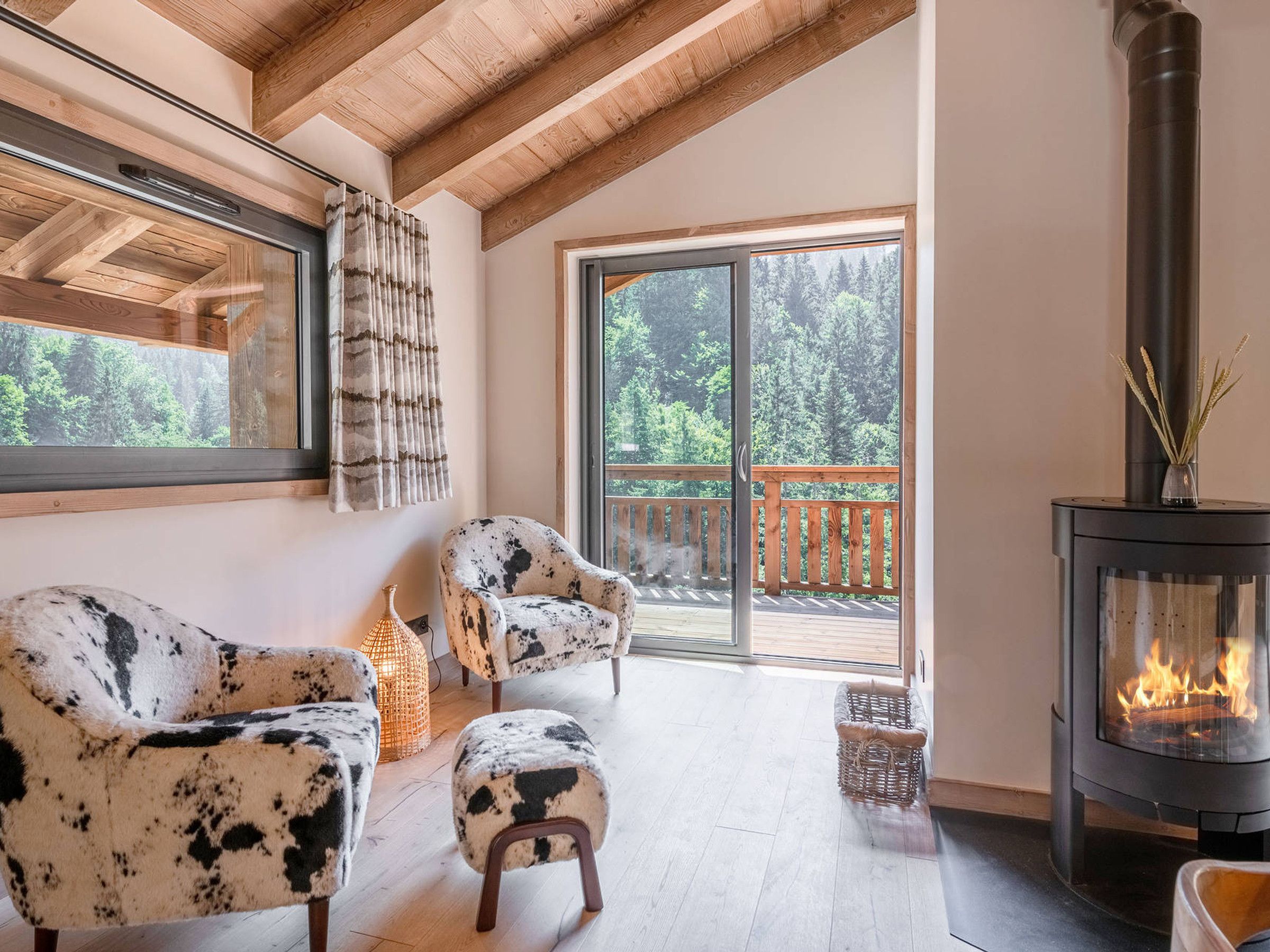 A cozy room with a fireplace, two patterned armchairs, a wicker basket, and a view of a balcony with a wooden railing.