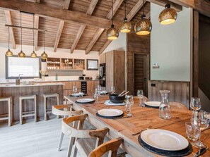 Une table de salle à manger en bois, dressée avec des assiettes, des verres et des couverts, dans une cuisine rustique aux poutres apparentes et dotée de luminaires suspendus.