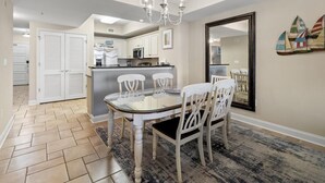 A dining area with a glass table and white chairs, a chandelier, and a kitchen in the background.