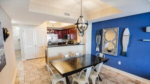A dining area with a black table and white chairs, a kitchen in the background, and a blue accent wall.