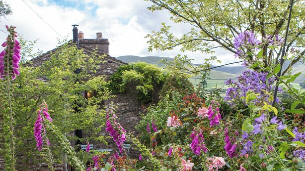 A stone house with a sloped roof, surrounded by lush greenery and flowering plants.