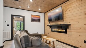 Un salon moderne avec un grand canapé d’angle, une cheminée en bois et un téléviseur à écran plat fixé au mur.