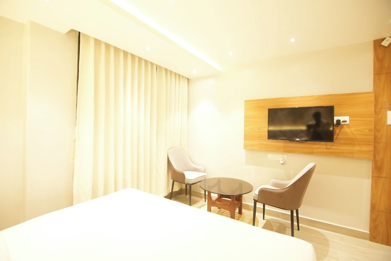 A modern hotel room with a large bed, two chairs, a small table, a TV mounted on the wall, and a large window with curtains.