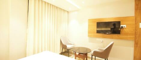 A modern hotel room with a large bed, two chairs, a small table, a TV mounted on the wall, and a large window with curtains.