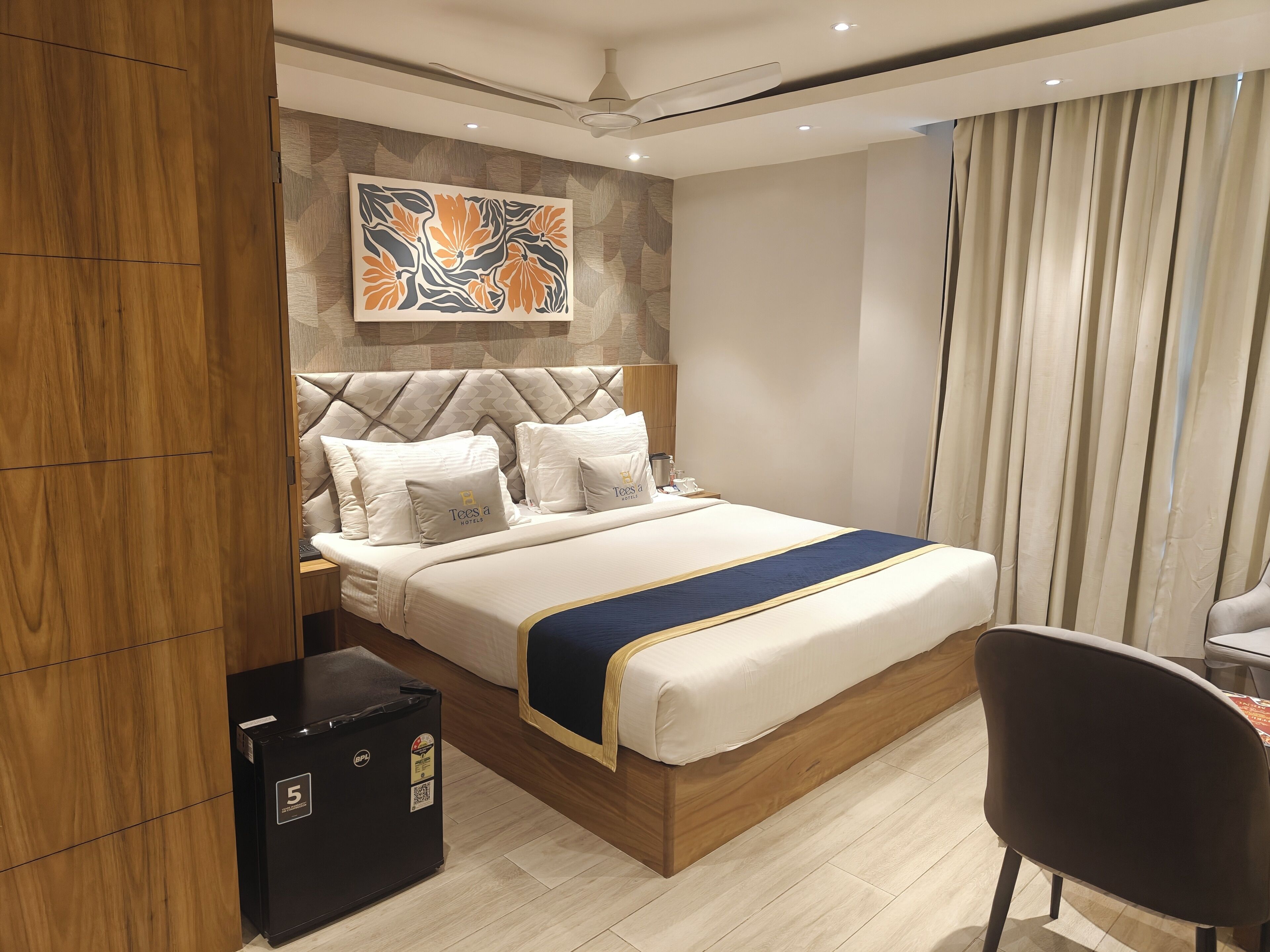 Ein Hotelzimmer mit einem Bett, einem Schreibtisch, einem Stuhl, einem kleinen schwarzen Kühlschrank und einer Wand mit einem Blumenbild.