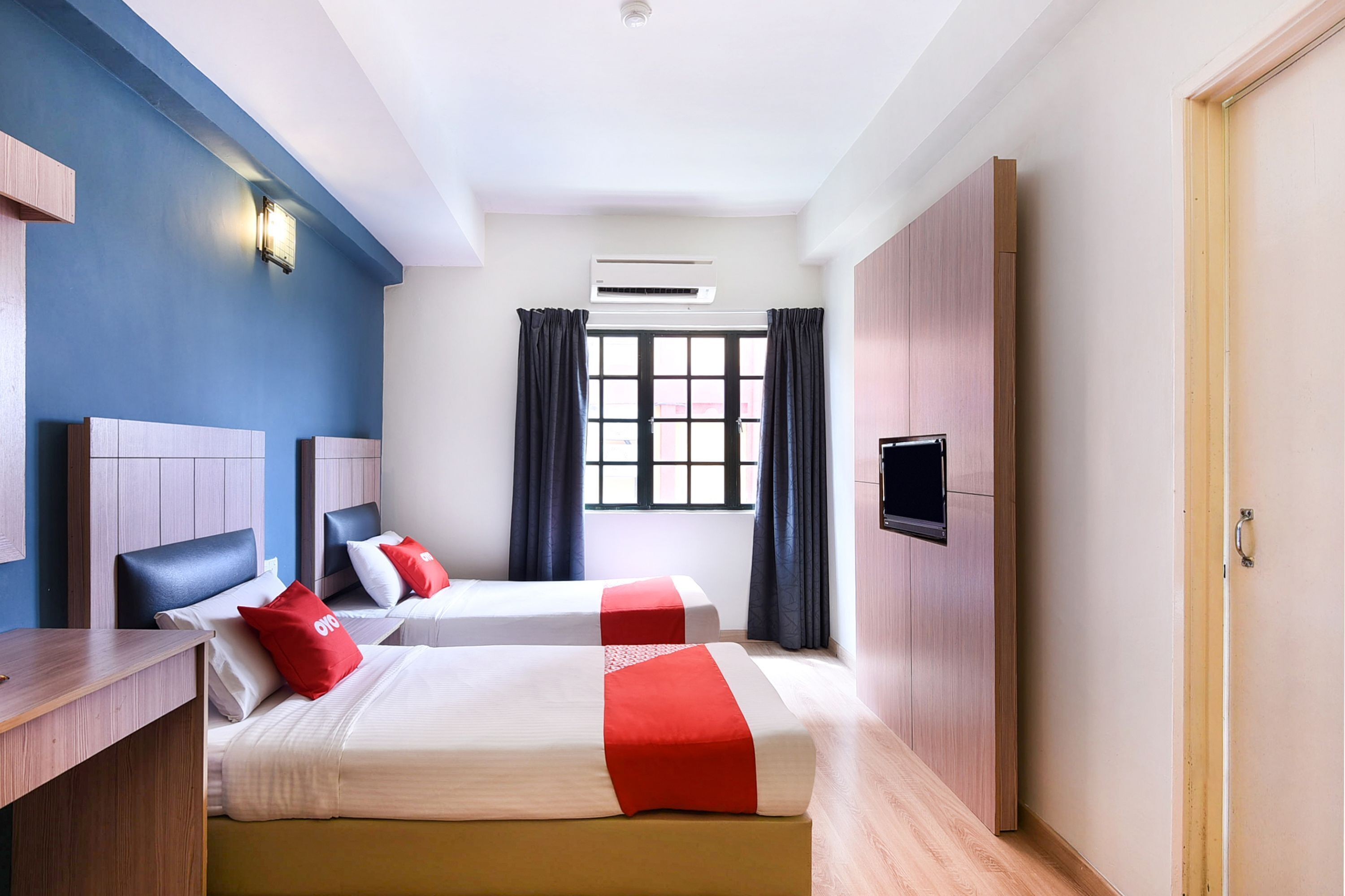 A hotel room with two beds, a TV, an air conditioning unit, and a window with curtains.