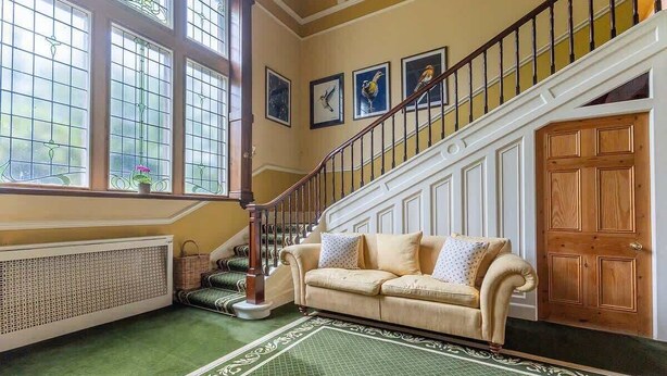 Een kamer met een groen tapijt met patroon, een beige bank met kussens, een houten deur en een trap met een leuning.