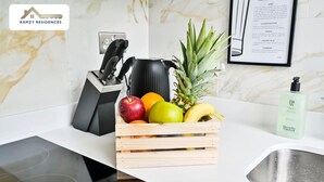 Una encimera de cocina con una cesta de frutas que contiene manzanas, naranjas y una piña. Hay un bloque de cuchillos, una botella de jabón para manos y un menú enmarcado en la pared.