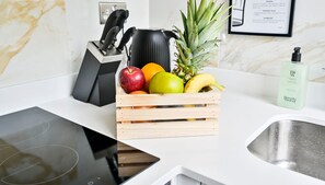 Una encimera de cocina con una cesta de frutas que contiene manzanas, naranjas y una piña. Hay un bloque de cuchillos, una botella de jabón para manos y un menú enmarcado en la pared.