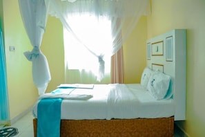 Ein Hotelzimmer mit Bett, einem Fenster mit Vorhängen, einem Nachttisch und einer Wand mit gerahmten Bildern.