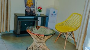 A room with a glass table, a wicker chair, a small potted plant, a painting, and a water dispenser. - The Tag Hotel  (Juja)
