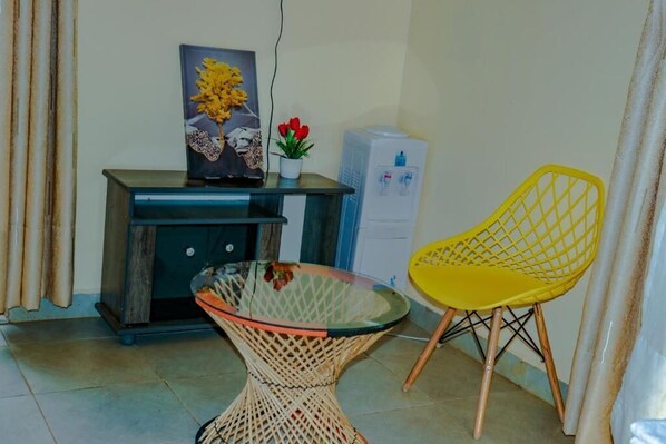 Quarto com mesa de vidro, cadeira de vime, pequena planta em vaso, pintura e dispensador de água.