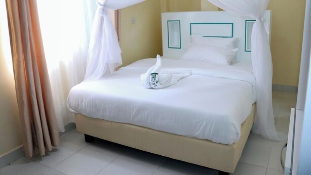 A neatly made bed with white bedding and a towel, a cream-colored headboard, and sheer white curtains. - The Tag Hotel  (Juja)