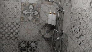 A shower area with a handheld showerhead and a wall-mounted faucet. The shower area has a tiled wall with various patterns and designs. There are two bottles of shampoo and conditioner on the wall. - Stanze letterarie  (Catania)