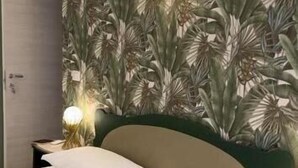 A neatly made bed with white linens, a green headboard, and a wall with a tropical leaf pattern. There are two bedside lamps with round shades. - Stanze letterarie  (Catania)