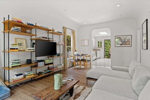 A modern living room with a flat-screen TV, wooden bookshelves, a grey sofa, and a dining area with a table and chairs.