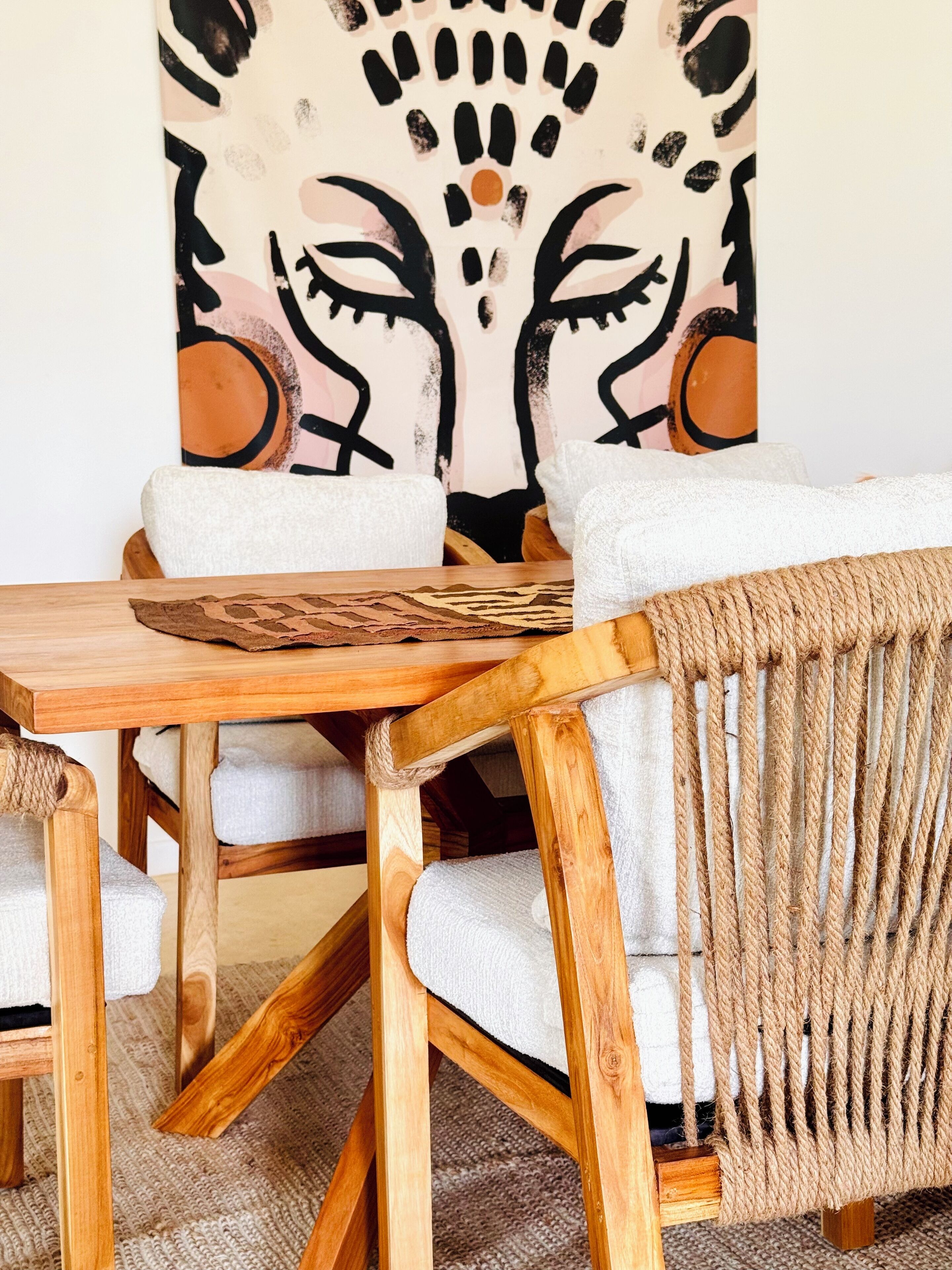 Una mesa de comedor de madera con una silla trenzada y una silla blanca acolchada. Un cuadro abstracto de gran tamaño adorna la pared detrás de la mesa.