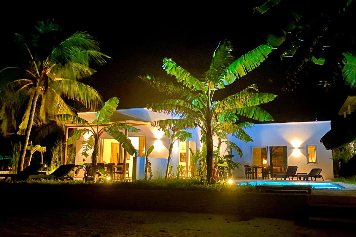 Propiedad frente a la playa de noche con piscina, palmeras y un edificio bien iluminado.