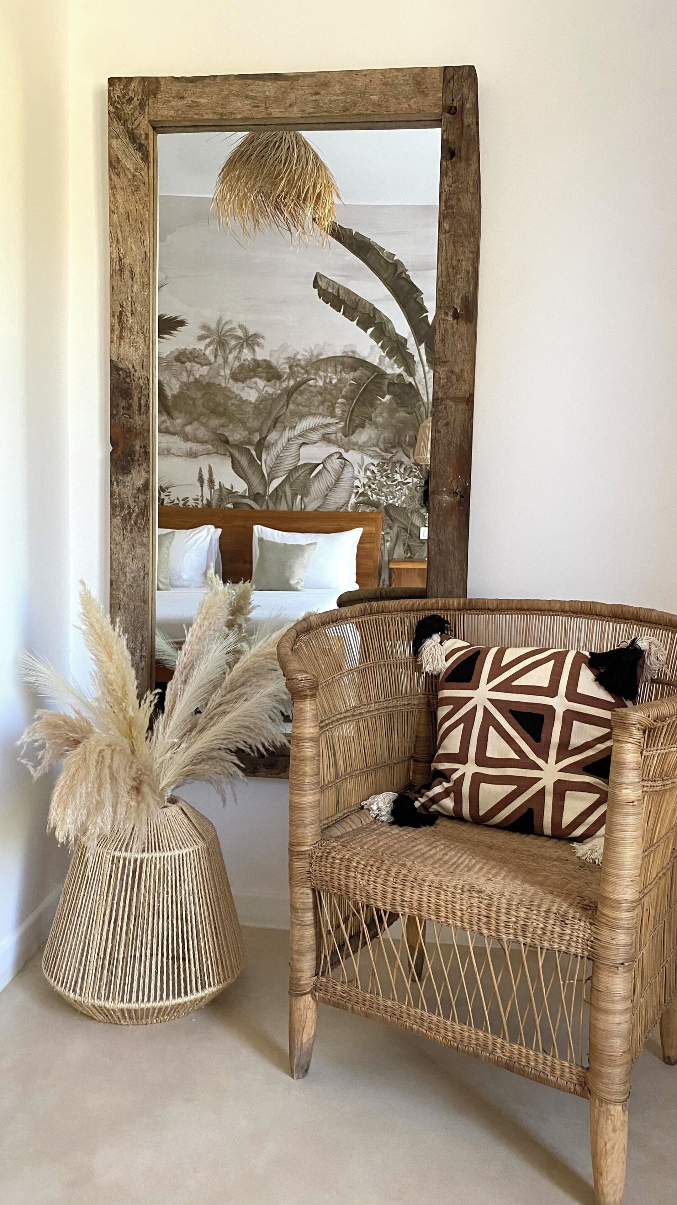 A wicker sofa with a Union Jack pillow, a woven basket with pampas grass, and a rustic mirror with a tropical landscape.