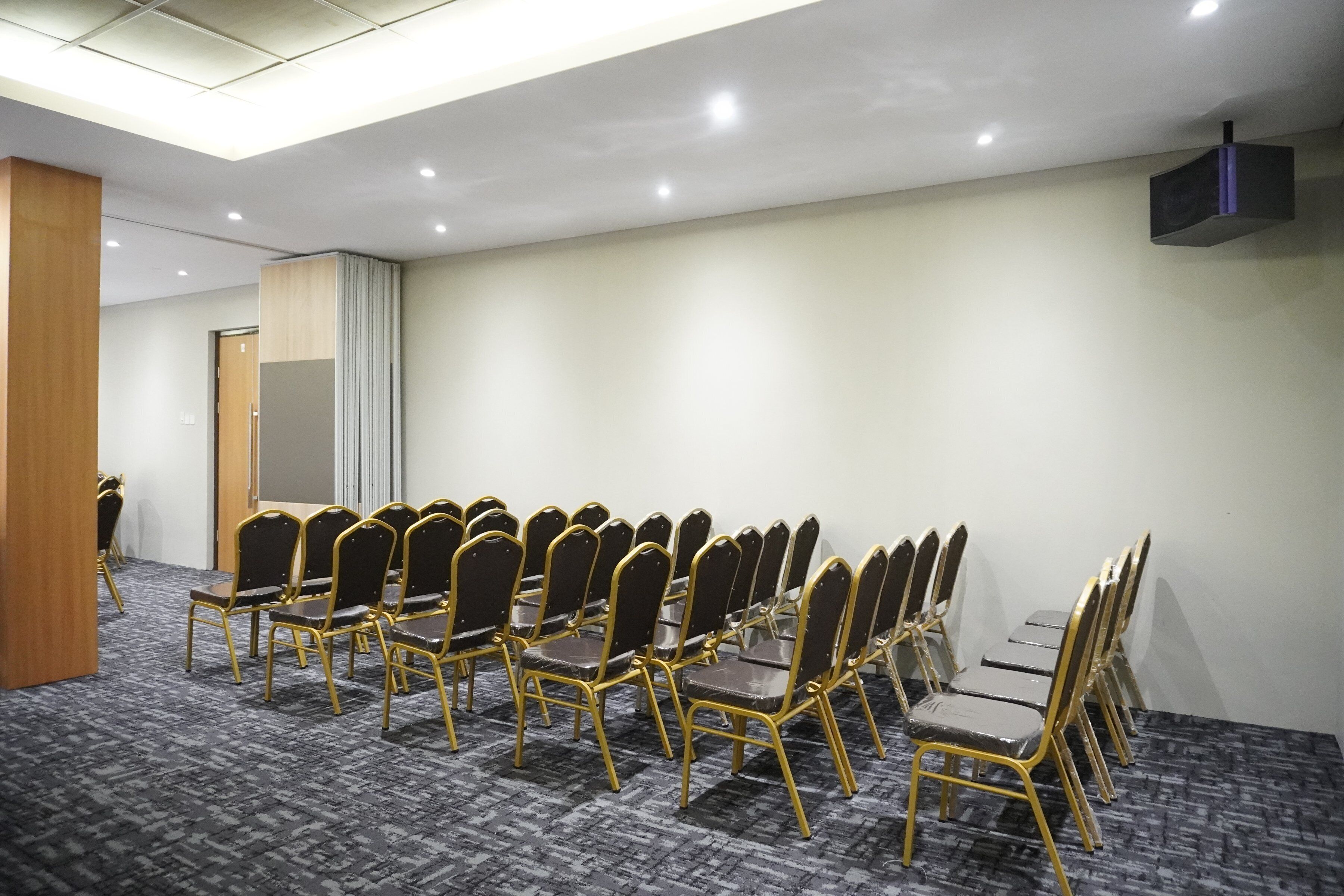 A conference room with rows of black chairs with gold frames, a projector mounted on the ceiling, and a carpeted floor.