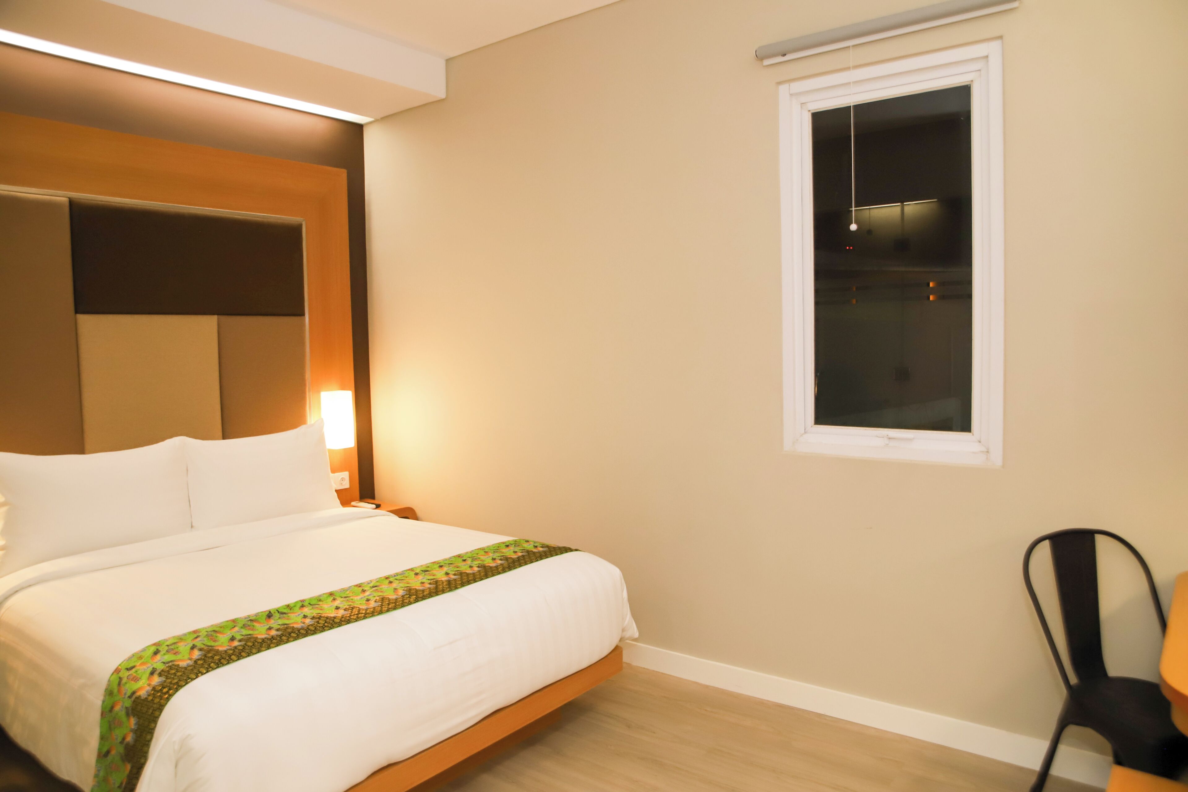A neatly made bed with a patterned bedspread, a bedside lamp, and a window with blinds.