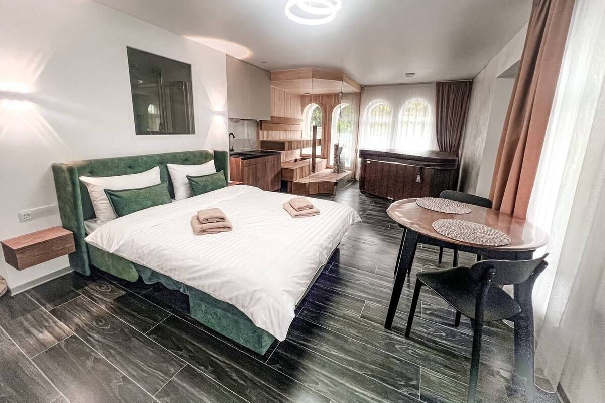 A modern hotel room with a large bed, a dining table, and a kitchen area.