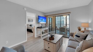A modern living room with a flat-screen TV, a sofa, a coffee table, and a sliding glass door leading to a balcony. - 2B/2B Fairview Isles 222 Condo, Walk to Beach, Heated Pool! (Fort Myers Beach)