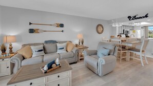 A modern living room with a sofa, armchairs, a coffee table, and a dining area with a table and chairs. There are decorative items like a lamp, a wall clock, and a sign that reads "Beach". - 2B/2B Fairview Isles 222 Condo, Walk to Beach, Heated Pool! (Fort Myers Beach)