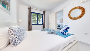 Un lit bien fait, orné d’un oreiller à motifs bleus et blancs, d’une serviette bleue et d’un tapis de bain bleu. Il y a une fenêtre avec des rideaux, un miroir au mur et une porte qui donne sur une autre pièce.