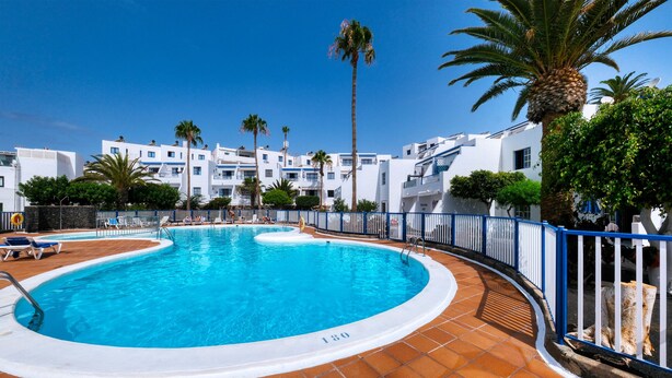 Une piscine dotée d’une terrasse carrelée, entourée de bâtiments blancs et de palmiers.