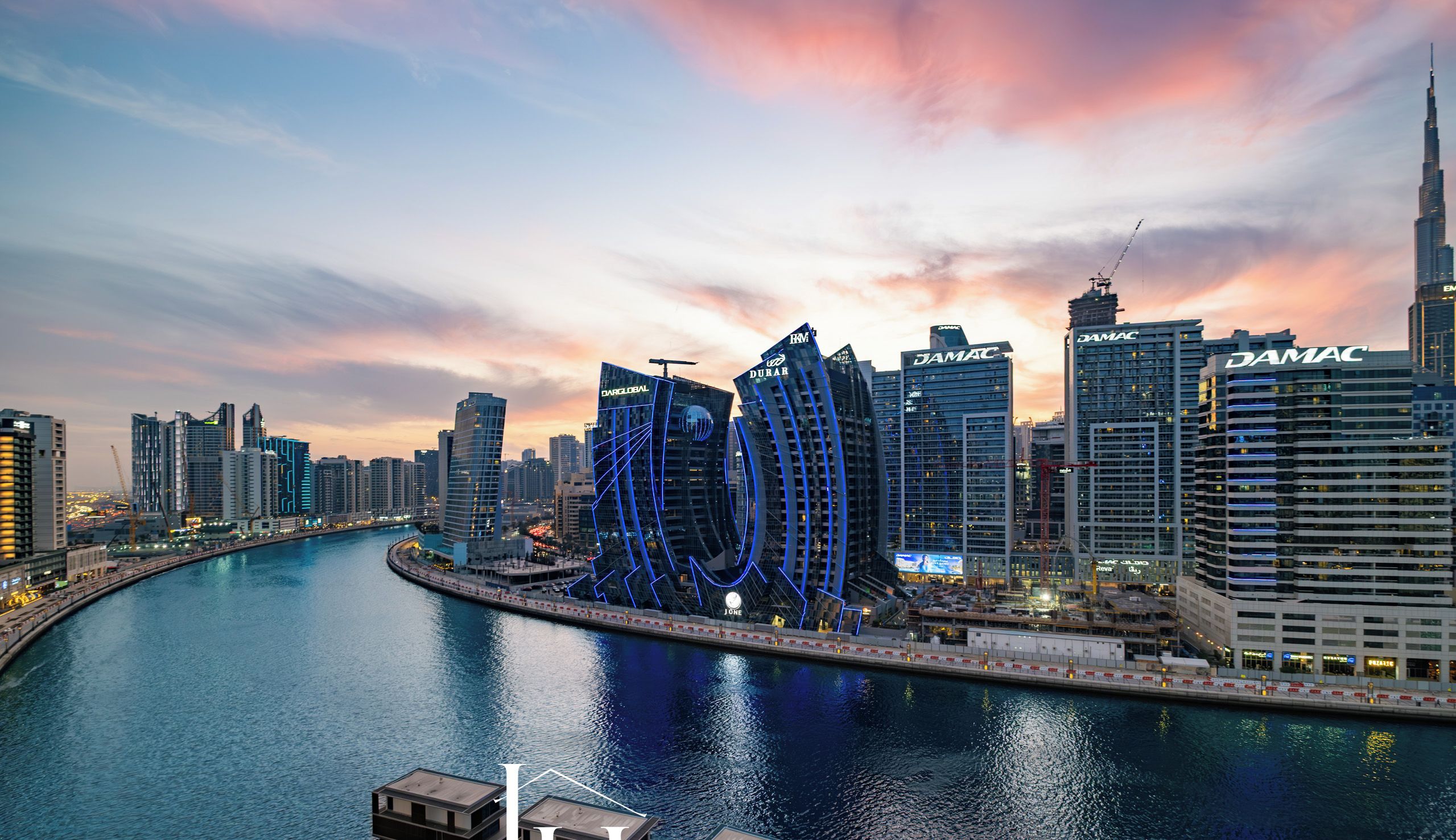 A waterfront cityscape with modern buildings, including a distinctive curved structure with illuminated blue lines, and the text "LUX HOLIDAY HOME DUBAI" at the bottom.