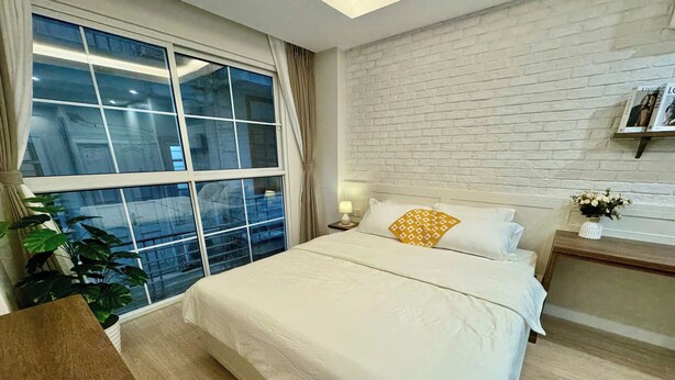 Ein Hotelzimmer mit großem Fenster, einem Bett mit weißer Bettwäsche und einem Zierkissen, einem Nachttisch mit Lampe und einer kleinen Pflanze.