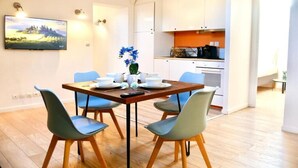 Zona pranzo con tavolo in legno e quattro sedie, televisore a parete e cucina sullo sfondo.