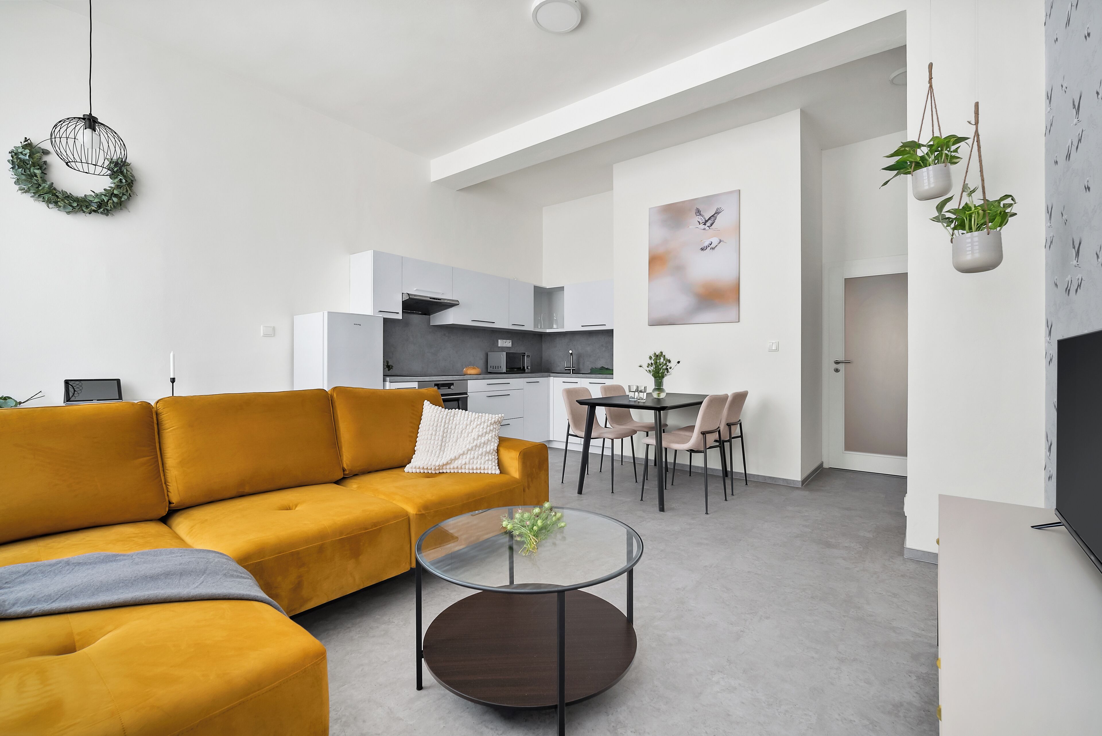 Een moderne woonkamer met een mosterdgele bank, een glazen salontafel en een keuken op de achtergrond.