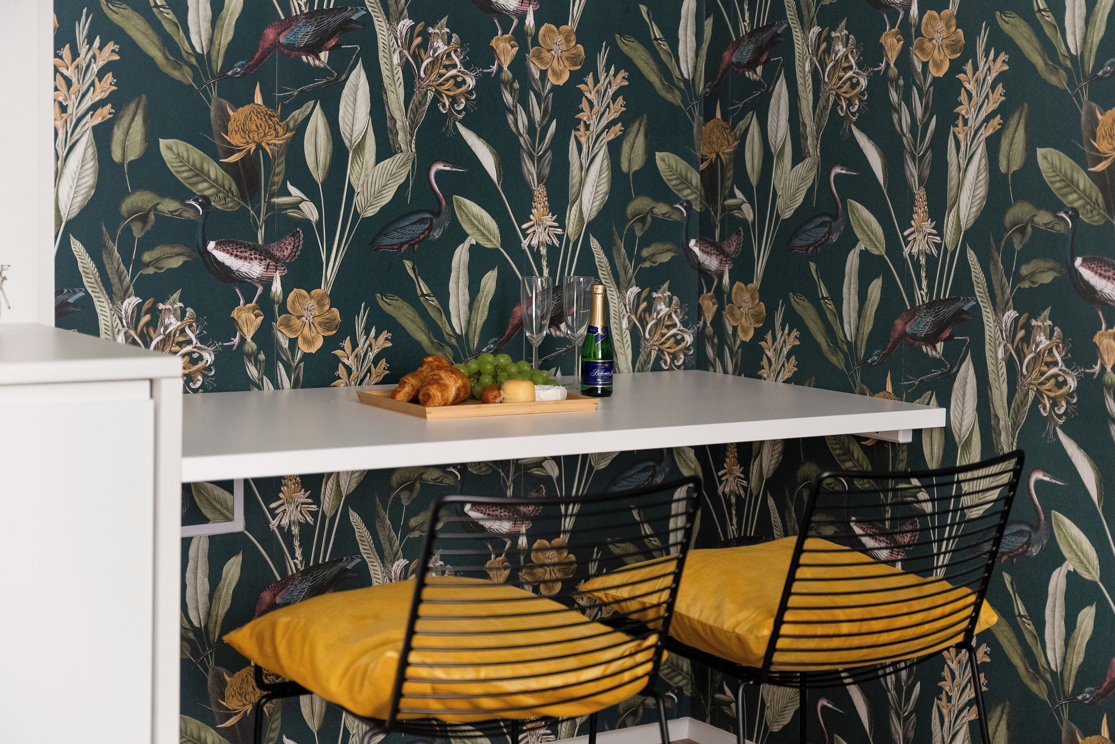 A dining area with a table set for two, featuring a floral wallpaper, a bottle of champagne, and a fruit bowl.