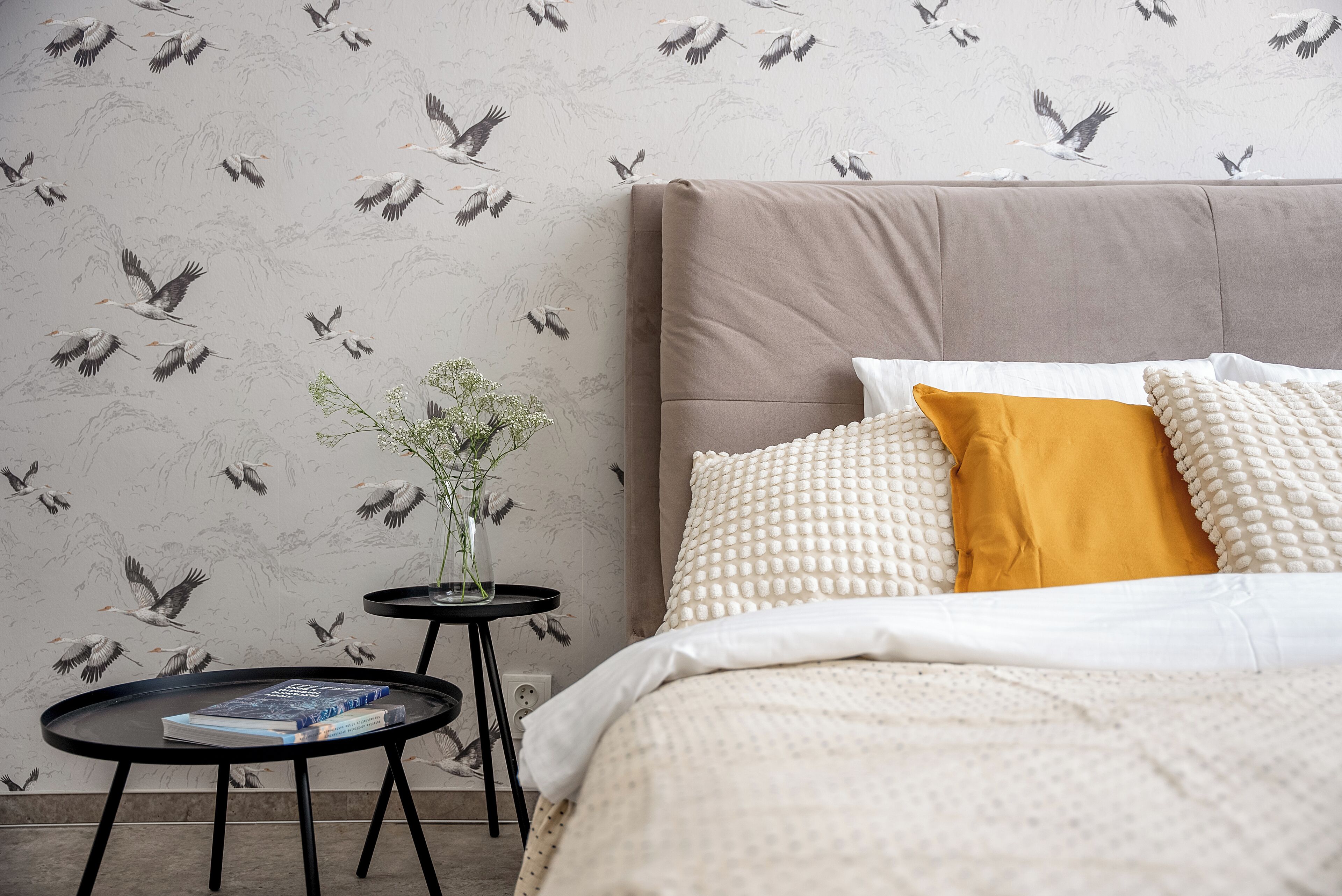 Ein Schlafzimmer mit einem großen Bett, einem Nachttisch mit einem Buch, einer Vase mit Blumen und einer Wand mit Vogelmotiv.