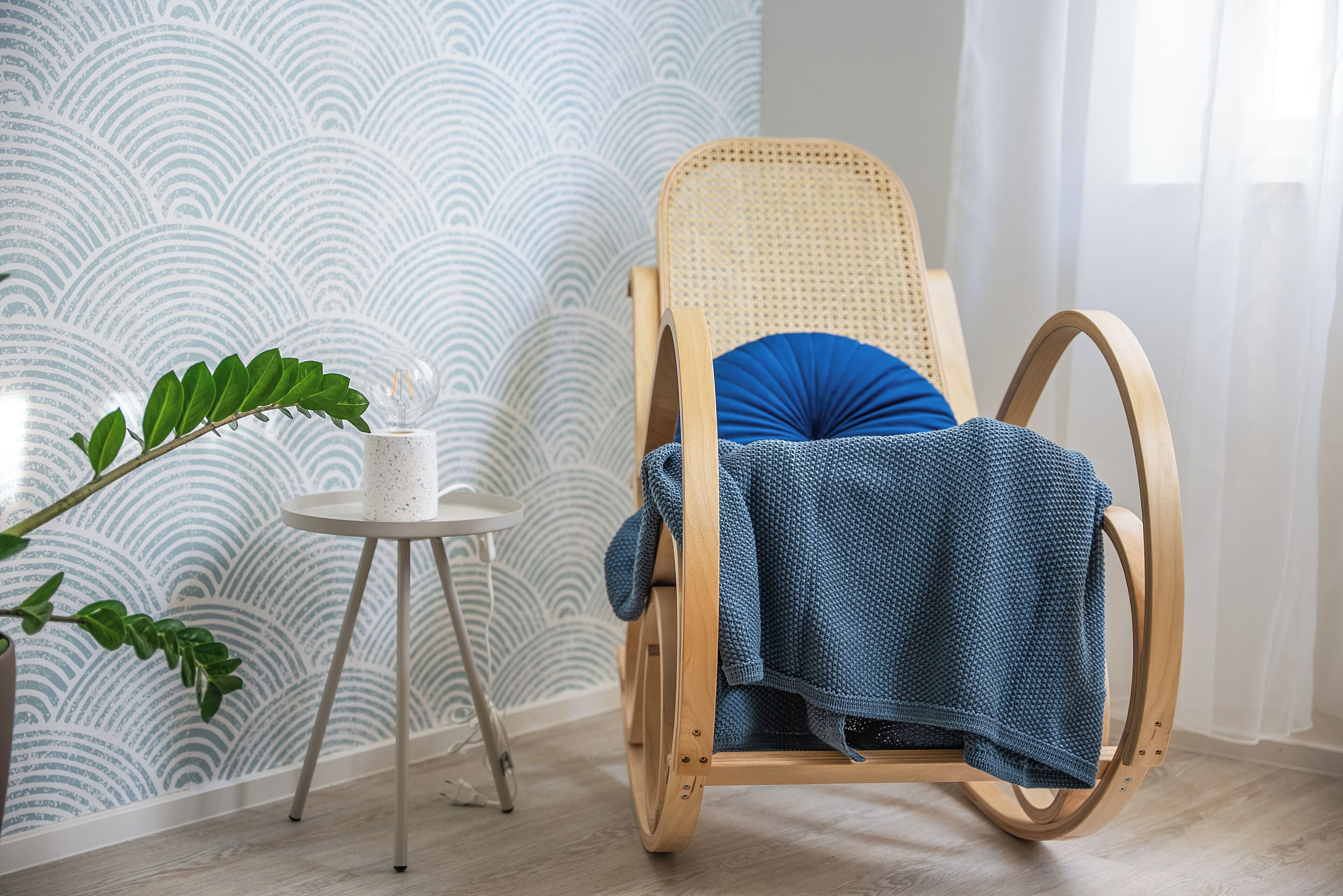 A wooden rocking chair with a blue cushion, a small white table with a plant, and a patterned wallpaper in a room with light wooden flooring.