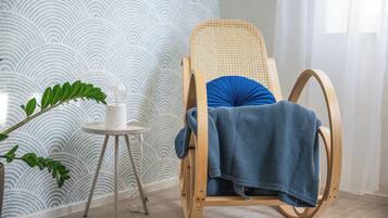 A wooden rocking chair with a blue cushion, a small white table with a plant, and a patterned wallpaper in a room with light wooden flooring.