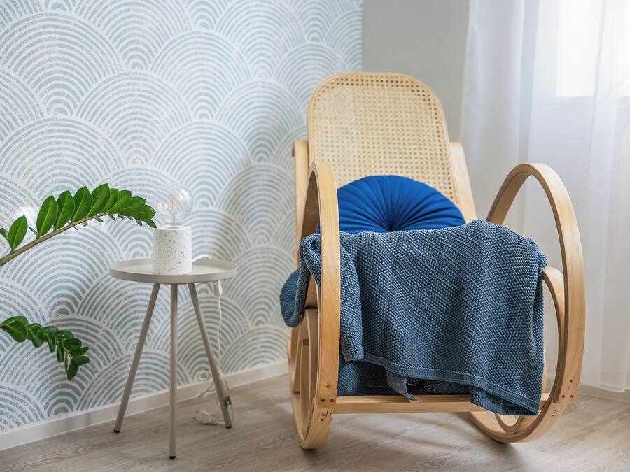 A wooden rocking chair with a blue cushion, a small white table with a plant, and a patterned wallpaper in a room with light wooden flooring.