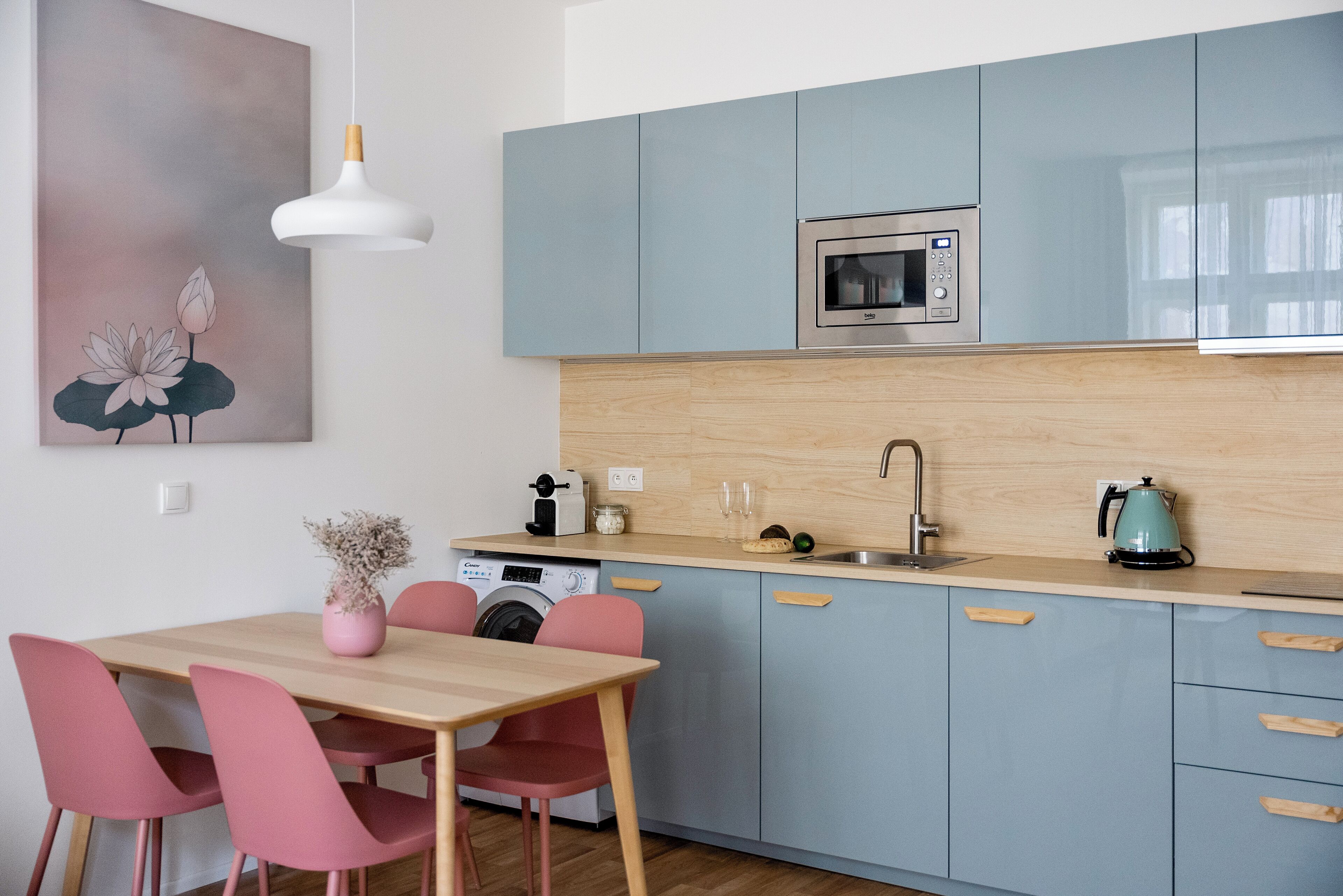 A modern kitchen with light blue cabinets, a wooden countertop, a built-in microwave, a sink, and a coffee maker. There is a dining table with pink chairs and a vase of flowers.